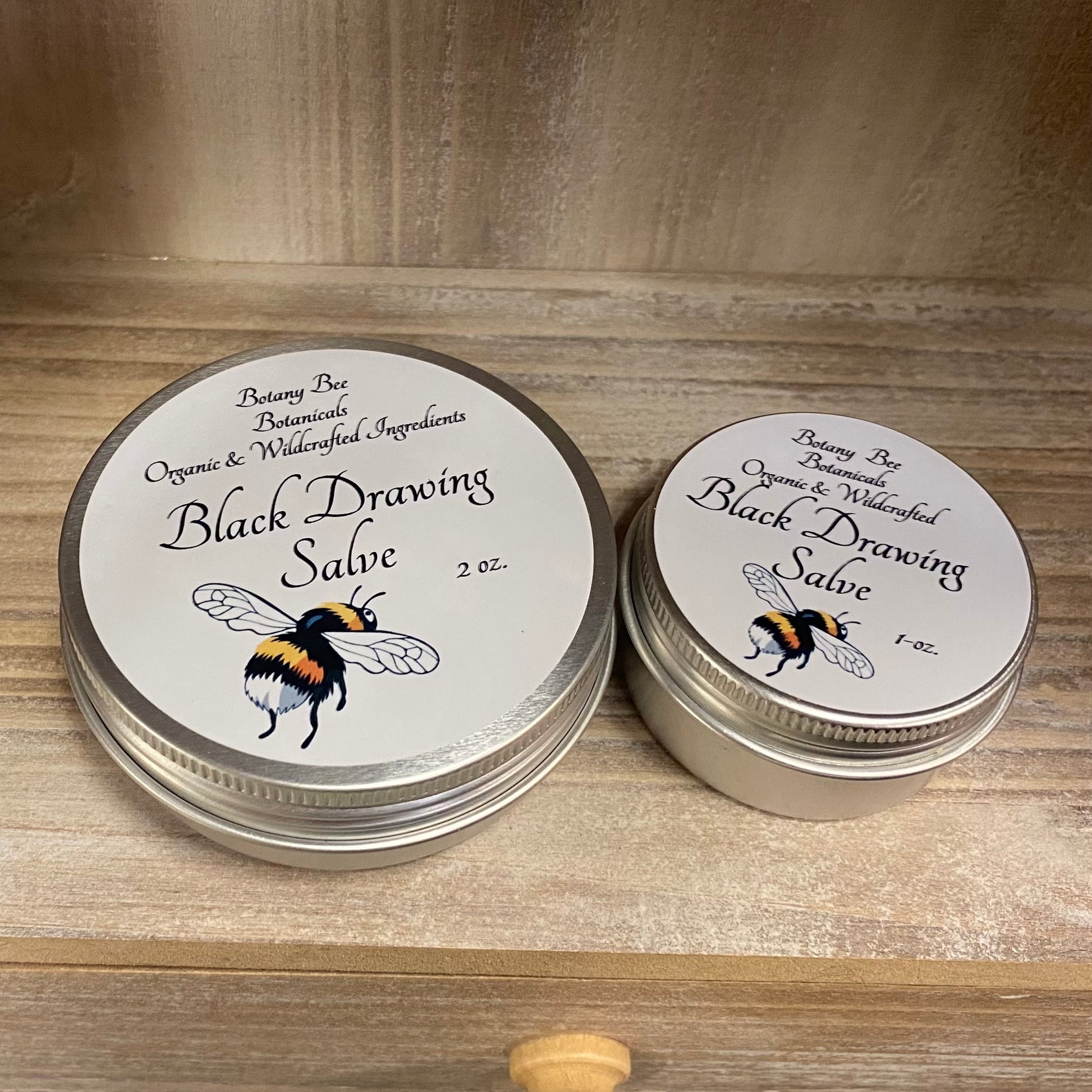 Two tins of Black Drawing Salve with labels on a wooden surface