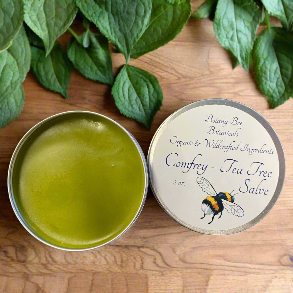 Open tin of green salve with 'Comfrey - Tea Tree Salve' label on a wooden surface