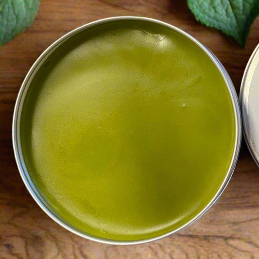 Open tin of green salve with 'Comfrey - Tea Tree Salve' label on a wooden surface