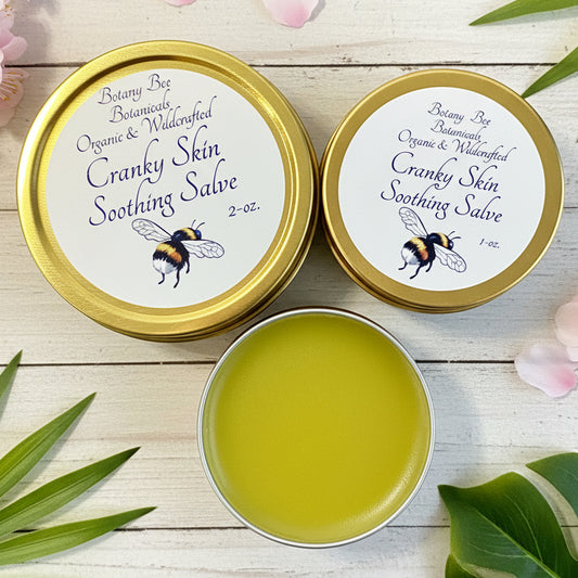 Two tins of 'Cranky Skin Soothing Salve' with a small open container on a wooden surface.