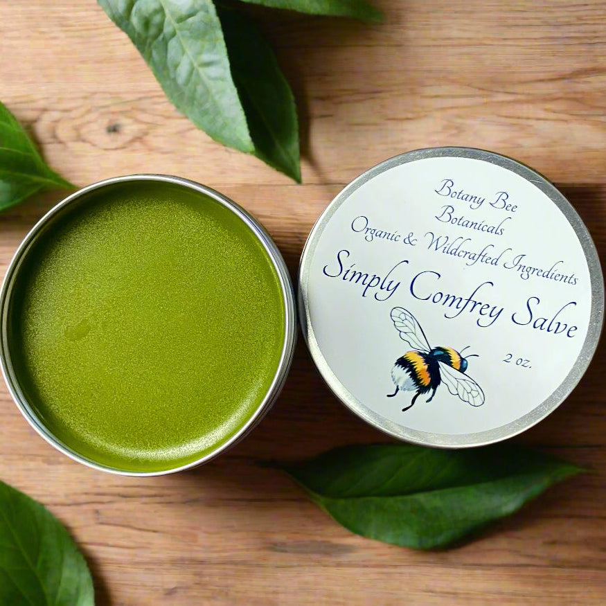 Open tin of green salve with Simply Comfrey Salve' label on a wooden surface with leaves.