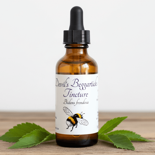 An amber glass dropper bottle with a black cap and a white label reading 'Devil's Bergartlaub Tincture' with a bumblebee illustration, set on a wooden surface with green leaves around it.