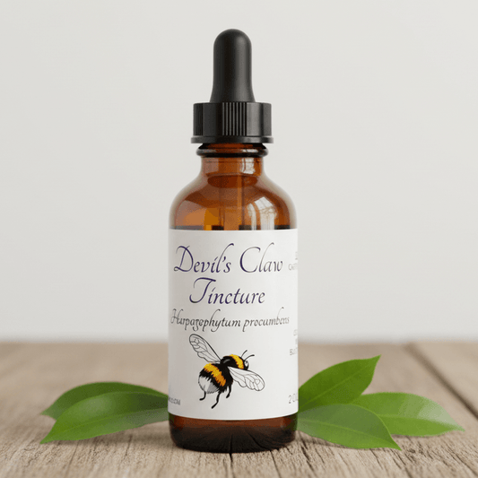 An amber glass dropper bottle with a black cap and a white label reading 'Devil's Claw Tincture' with a bee illustration, placed on a wooden surface with green leaves around it.