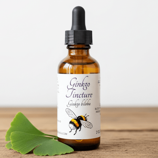 An amber glass dropper bottle labeled 'Ginkgo Tincture Ginkgo biloba' with a black dropper cap, standing on a wooden surface against a light background, with green ginkgo leaves placed beside it.