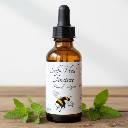 An amber glass dropper bottle labeled 'Self-Heal Tincture' with a black cap, placed on a wooden surface with green mint leaves around it. The label includes a bee illustration and the text 'Punica vulgaris'.